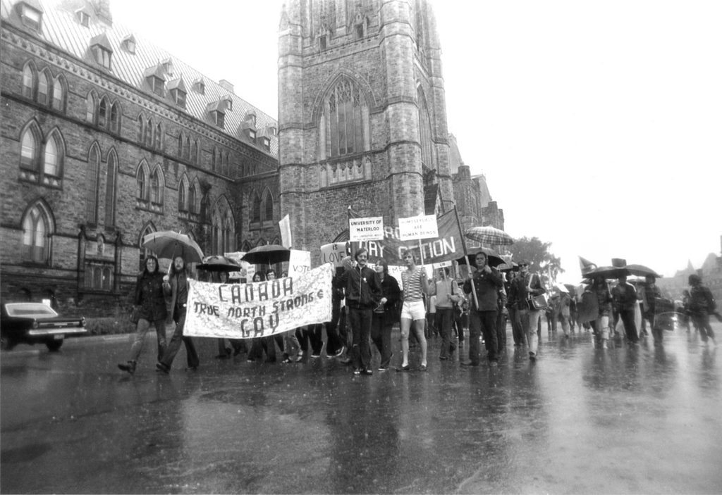 Une foule de manifestants défile devant la Colline du Parlement lors du rassemblement « We Demand » de 1971. Tenant des pancartes et des parapluies sous la pluie, les personnes à l’avant de la foule brandissent une bannière sur laquelle on peut lire : « Canada True North Strong & Gay » 